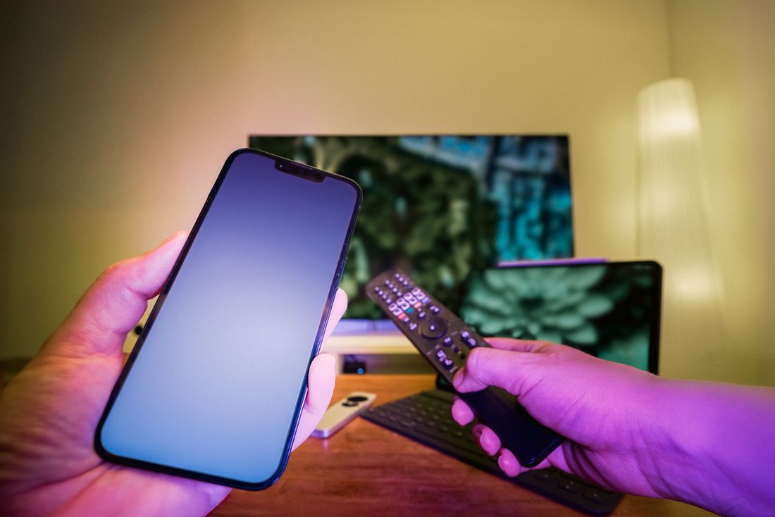 Hands holding a smartphone and television remote in a home media setup.
